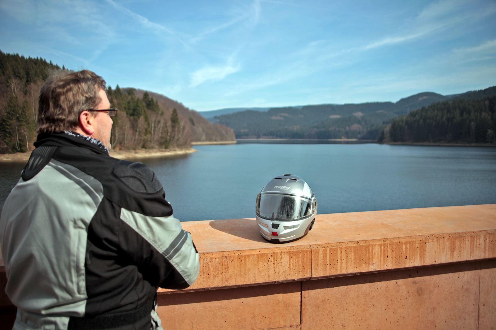 Mann in Motorradjacke an einer Mauerbrüstung, daneben ein silberner Helm, dahinter der Stausee Mann in Motorradjacke an einer Mauerbrüstung, daneben ein silberner Helm, dahinter der Stausee