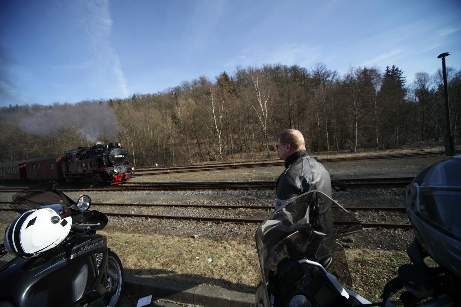 Mann in Lederkombi an mehreren Gleisen, links eine Dampflok mit rotem Wagen, vorn zwei Motorräder Mann in Lederkombi an mehreren Gleisen, links eine Dampflok mit rotem Wagen, vorn zwei Motorräder
