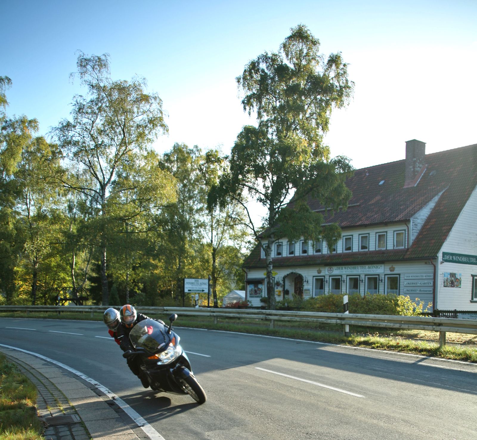 Motorrad mit Sozius in Schräglage in einer Linkskurve, rechts hinter der Leitplanke ein weißes Holzhaus mit Werbeschildern Motorrad mit Sozius in Schräglage in einer Linkskurve, rechts hinter der Leitplanke ein weißes Holzhaus mit Werbeschildern