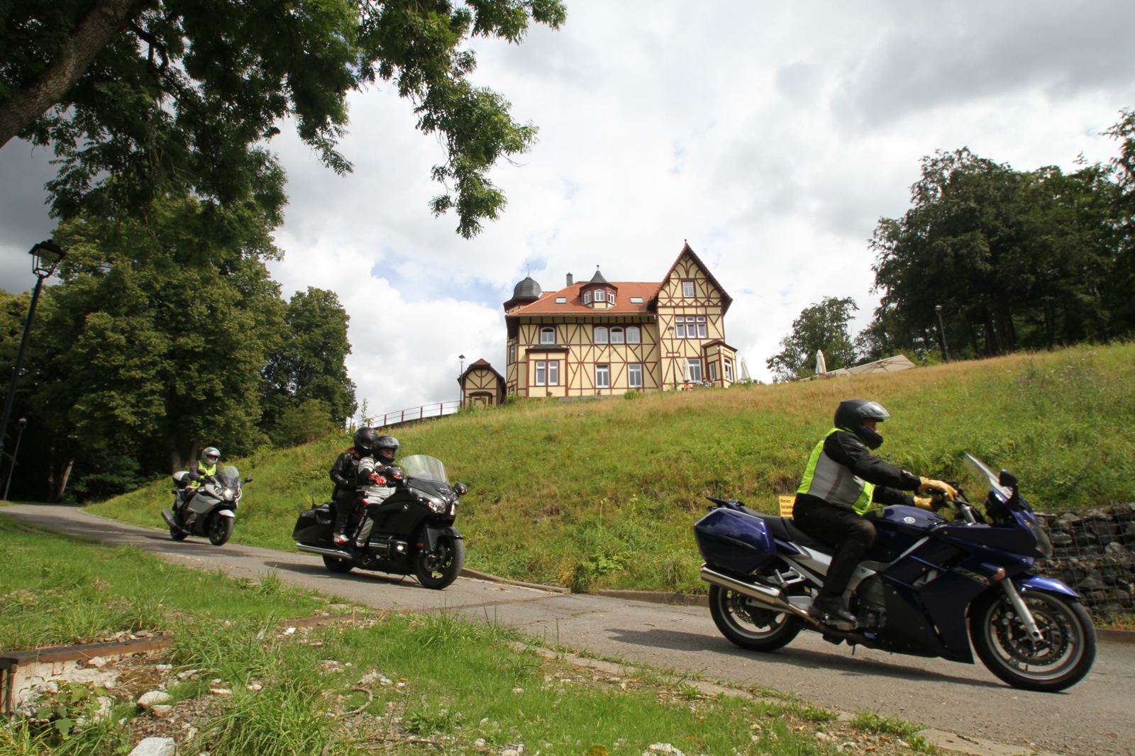 Drei Motorräder fahren eine schmale Straße hinunter, oberhalb ein großes Fachwerkhaus mit Turm Drei Motorräder fahren eine schmale Straße hinunter, oberhalb ein großes Fachwerkhaus mit Turm