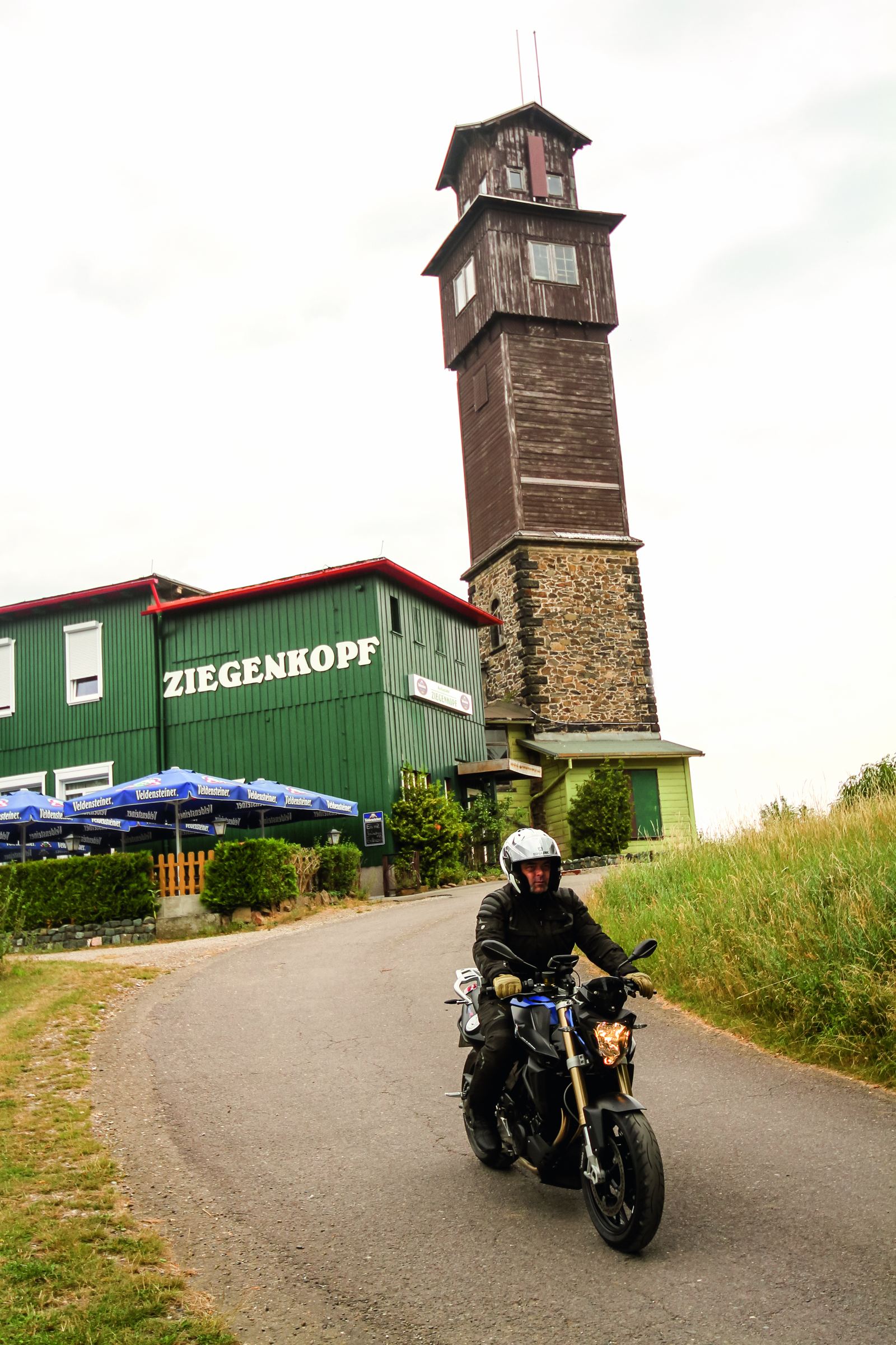 Motorradfahrer auf einer Zufahrt, dahinter ein grünes Gasthaus und ein hoher hölzerner Aussichtsturm Motorradfahrer auf einer Zufahrt, dahinter ein grünes Gasthaus und ein hoher hölzerner Aussichtsturm