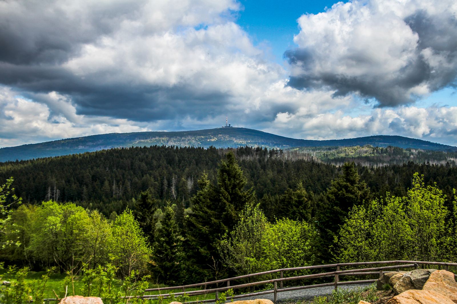 Bewaldeter Höhenzug unter Wolkenhimmel, dahinter eine flache Kuppe mit Sendemast, im Vordergrund ein Holzgeländer Bewaldeter Höhenzug unter Wolkenhimmel, dahinter eine flache Kuppe mit Sendemast, im Vordergrund ein Holzgeländer
